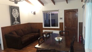A dining area with a wooden table, four chairs, a dark brown sofa, and a window with a view of the outside. - Cabañas Cerro Las Piedras (Punta Arenas)