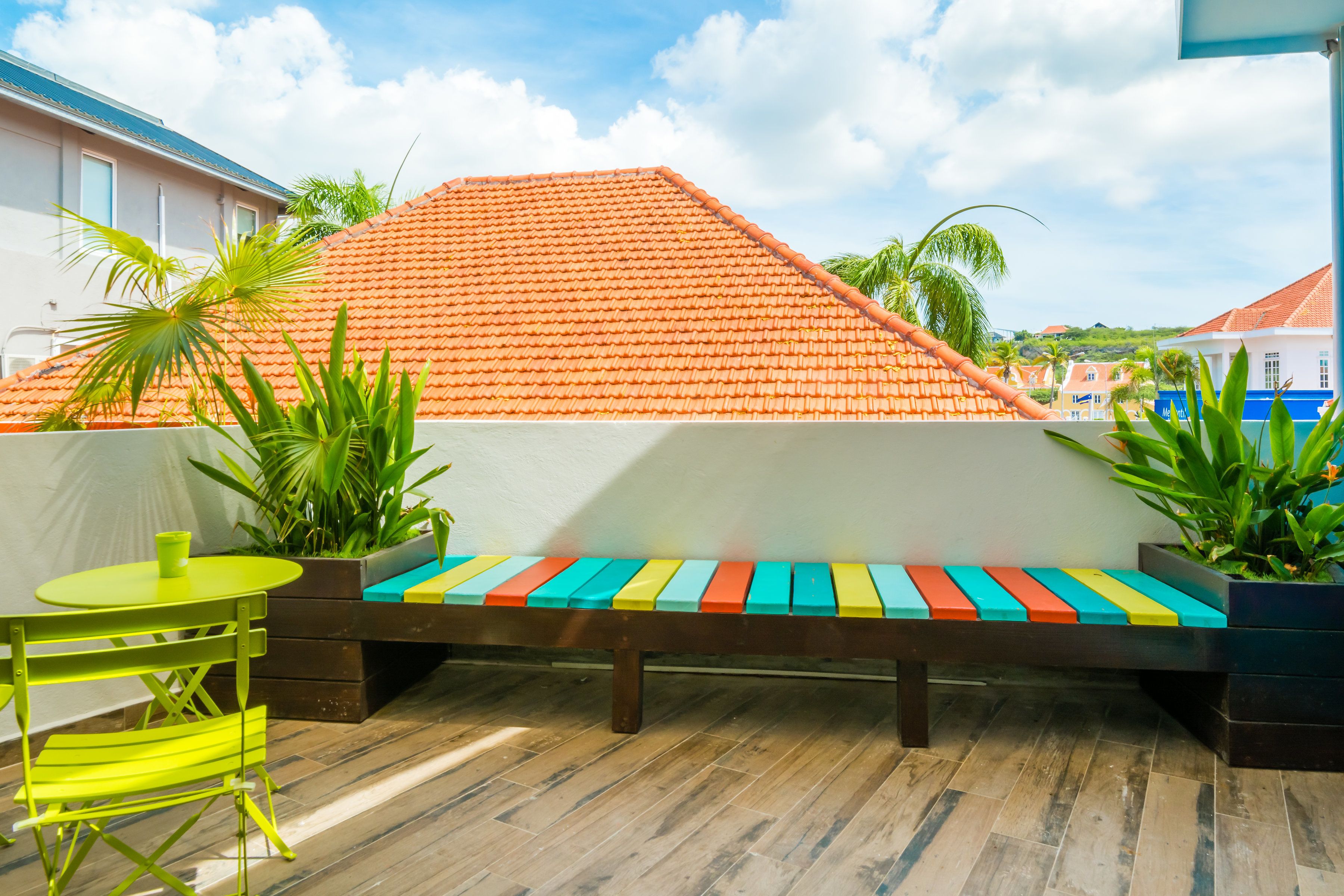Een terras met een houten bank, een gele tafel en groene stoelen.