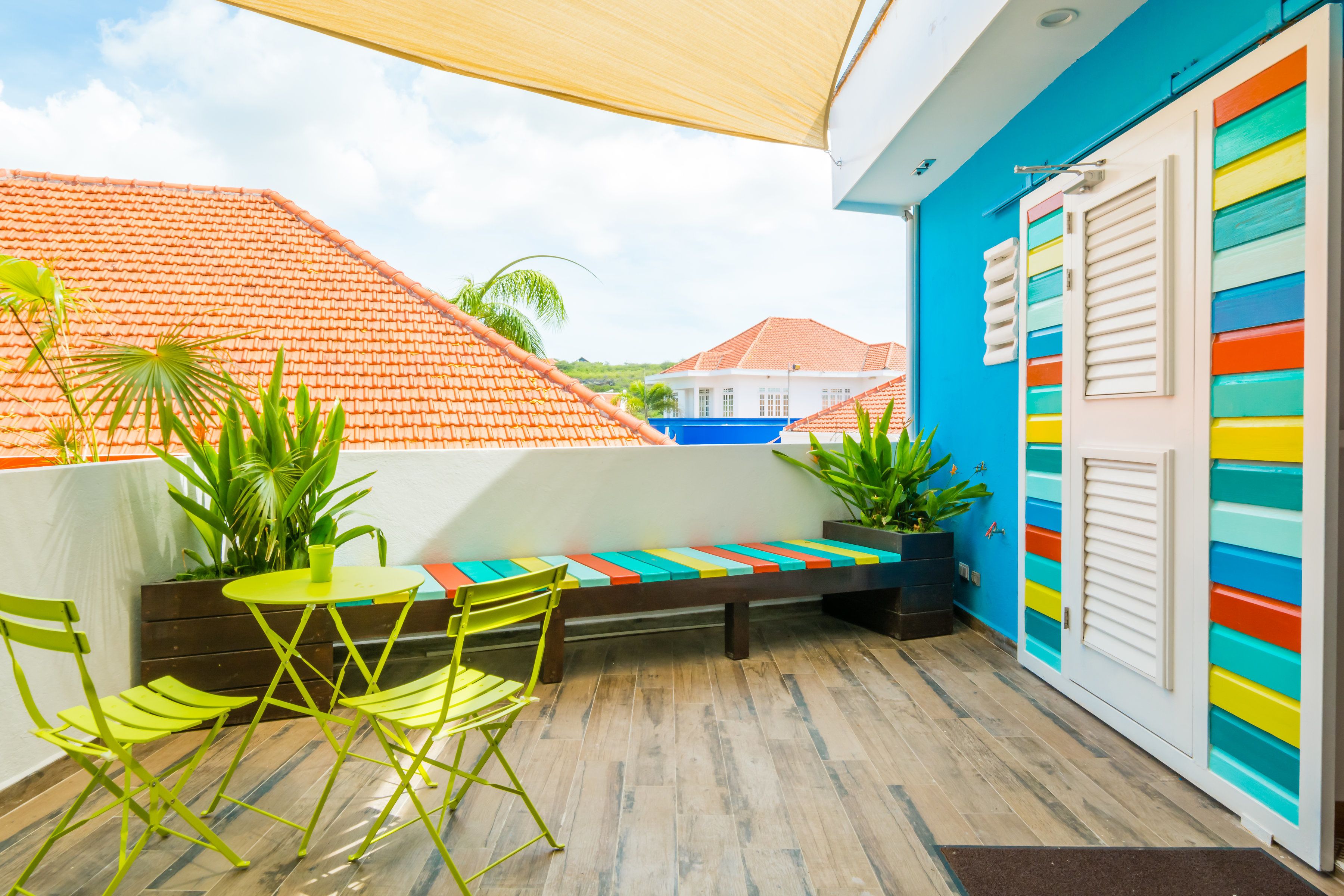 A balcony with a wooden floor, a green folding chair, a small round table, and a colorful bench.