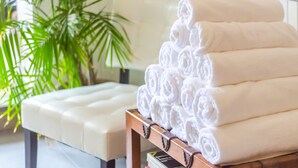 A stack of neatly rolled white towels on a wooden bench in a room with a white cushioned bench and a green plant in the background. - Bed & Bike Curacao Hostel (Willemstad)