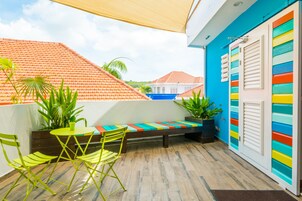A balcony with a wooden floor, a green folding chair, a small round table, and a colorful bench.