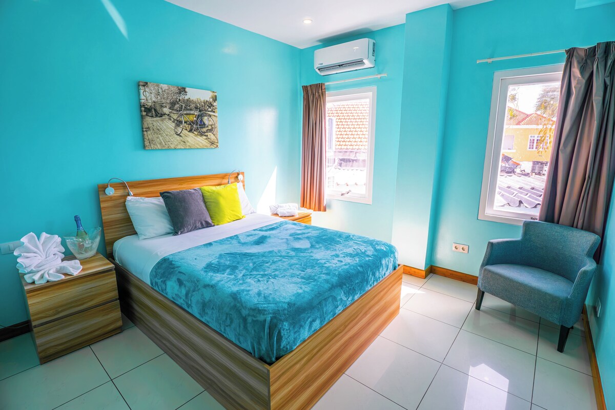 Une chambre d’hôtel comprenant un lit, une table de chevet, une chaise, une fenêtre avec des rideaux et un appareil de climatisation.