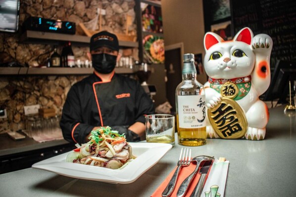 Un chef prépare un plat dans un restaurant. On peut voir une statue de chat décorative et une bouteille d’alcool sur le comptoir.