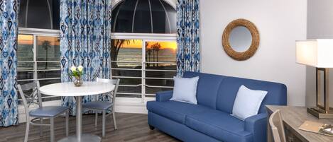 A room with a blue sofa, a round table with chairs, and a view of the sunset through large windows with patterned curtains.