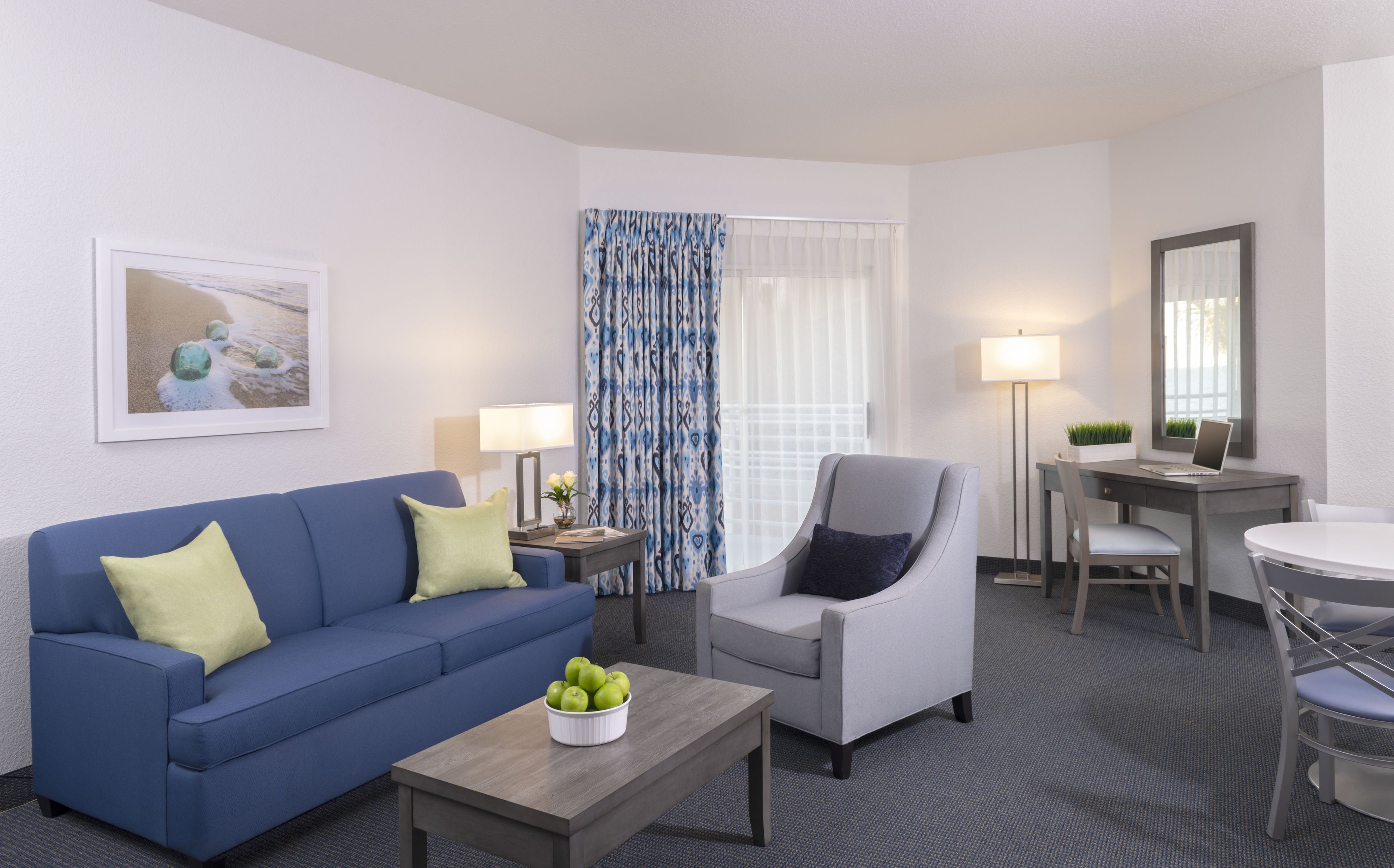 Una habitación de hotel moderna con un sofá azul, un sillón gris, una mesa de centro de madera con manzanas verdes, un escritorio con una computadora portátil y un ventanal con cortinas estampadas.