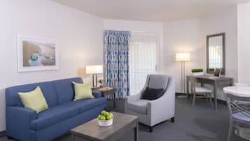 Una habitación de hotel moderna con un sofá azul, un sillón gris, una mesa de centro de madera con manzanas verdes, un escritorio con una computadora portátil y un ventanal con cortinas estampadas.