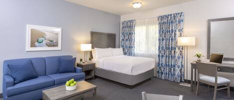 A hotel room with a bed, a blue sofa, a desk with a chair, a small table with fruit, and a lamp.