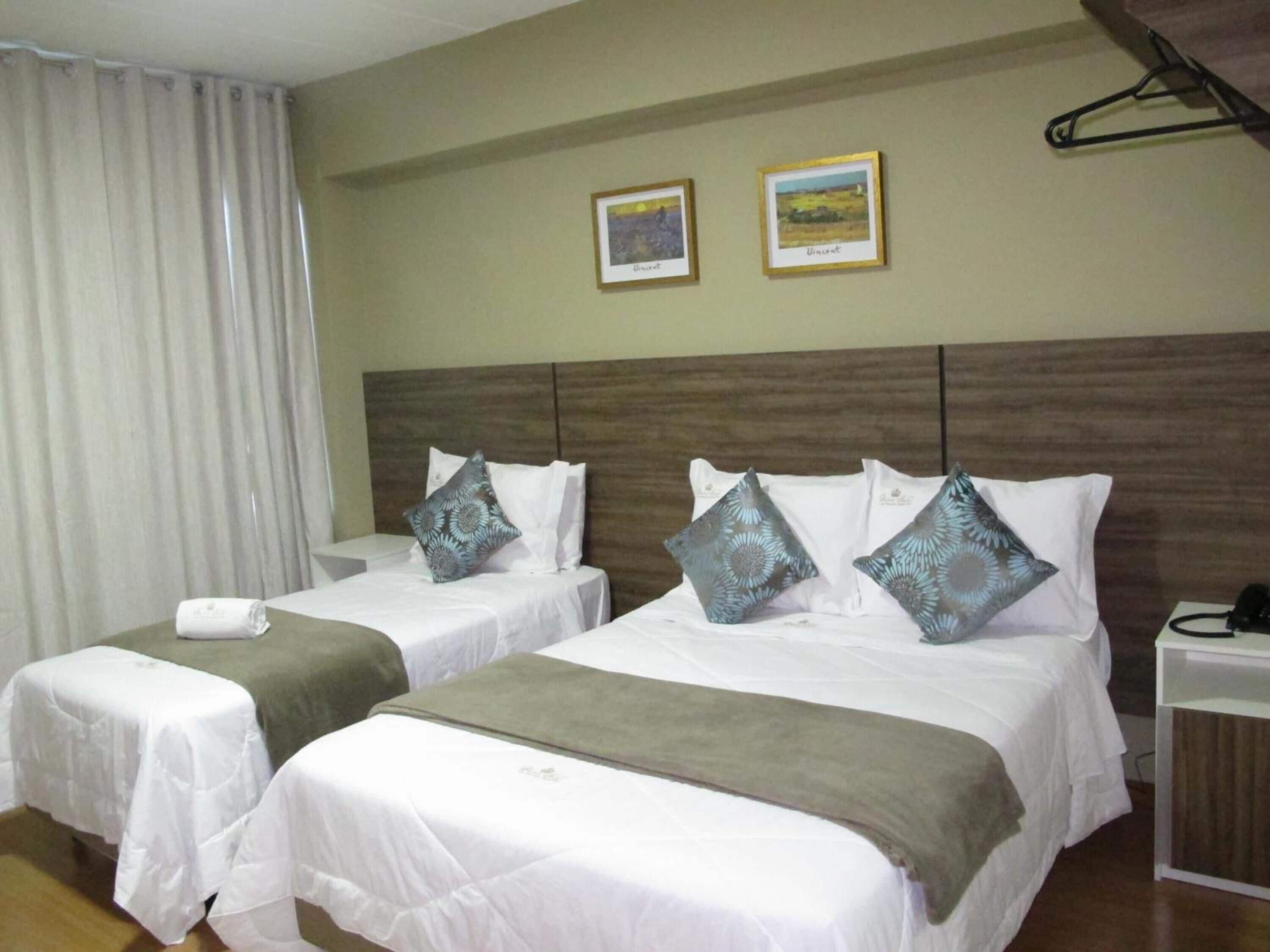 A hotel room with two beds, each with a pillow and a blanket. There is a nightstand with a phone and a lamp. The room has a wooden headboard and a curtain on the window.