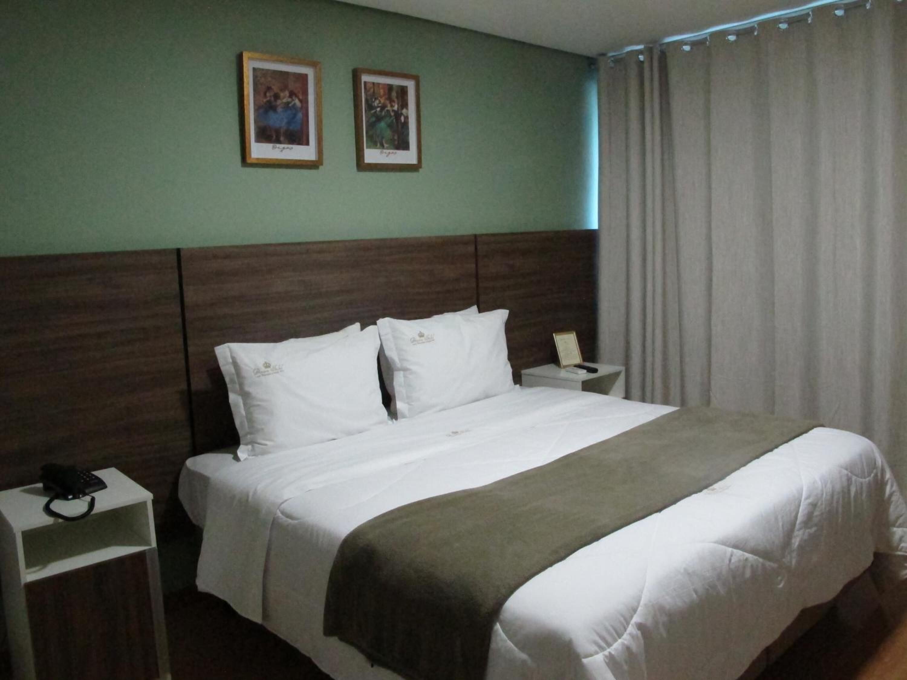Une chambre d’hôtel avec un grand lit, une tête de lit en bois, deux tables de chevet, une petite table et un rideau.