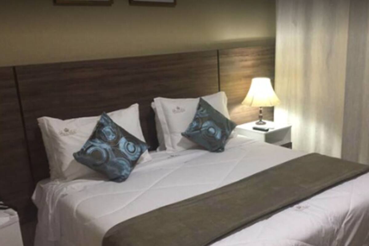 A hotel room with a large bed, white bedding, two decorative pillows, a bedside lamp, and a small table.
