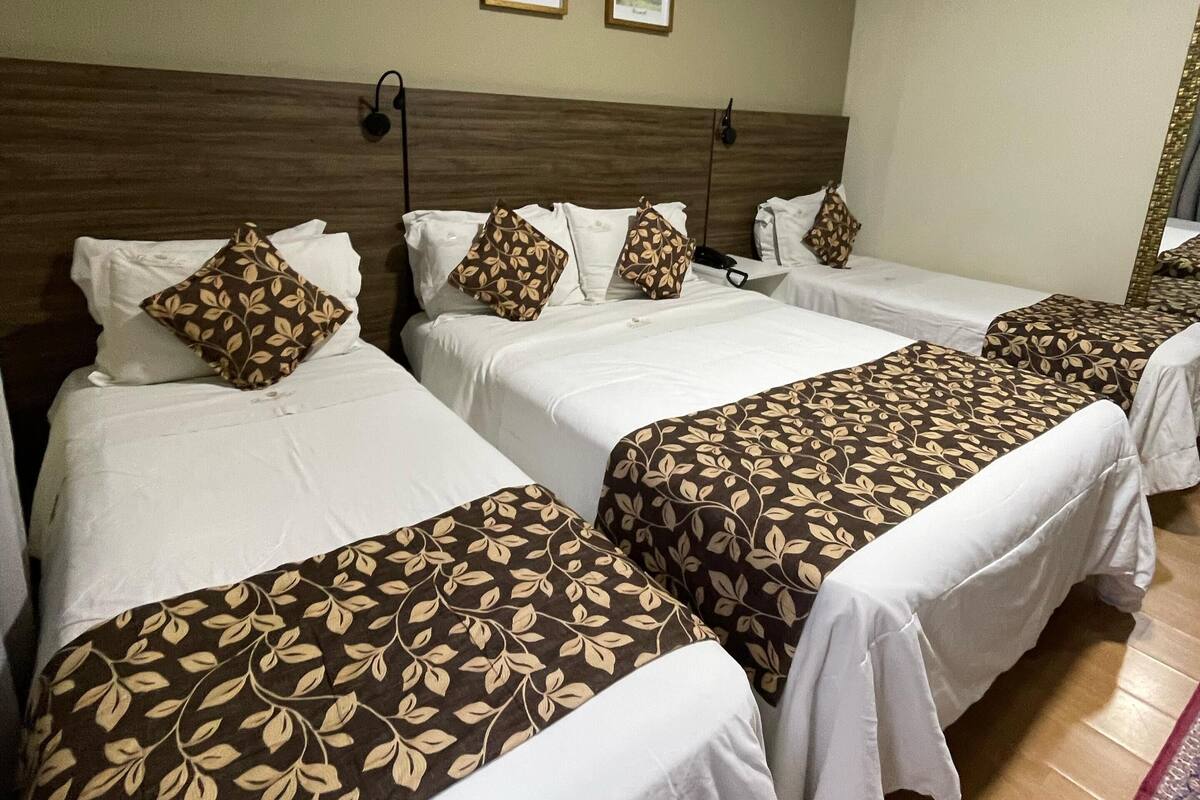 A hotel room with four single beds, each with a patterned bedspread and matching pillows. There are two framed pictures on the wall above the beds, and an air conditioning unit mounted on the wall.