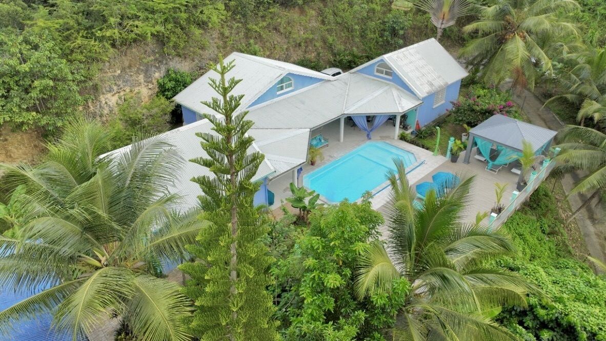 Una casa de dos pisos con piscina, rodeada de exuberante follaje y palmeras.