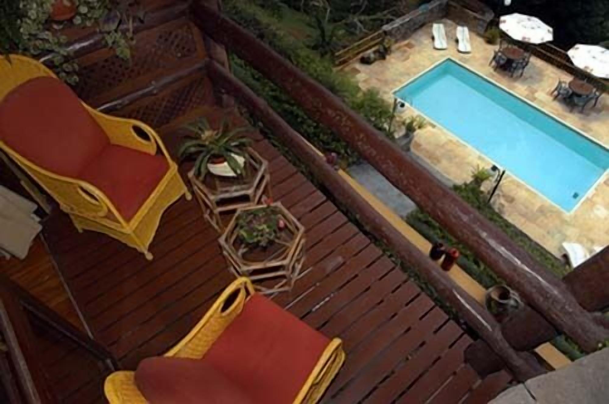 A wooden deck with two red chairs, a small table with a plant, and a view of a swimming pool with lounge chairs and umbrellas.