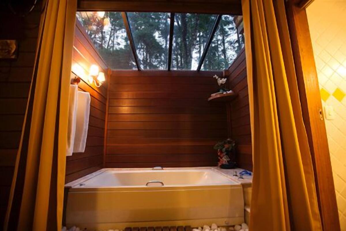 A wooden cabin with a large bathtub, a window with curtains, and a small shelf with items.