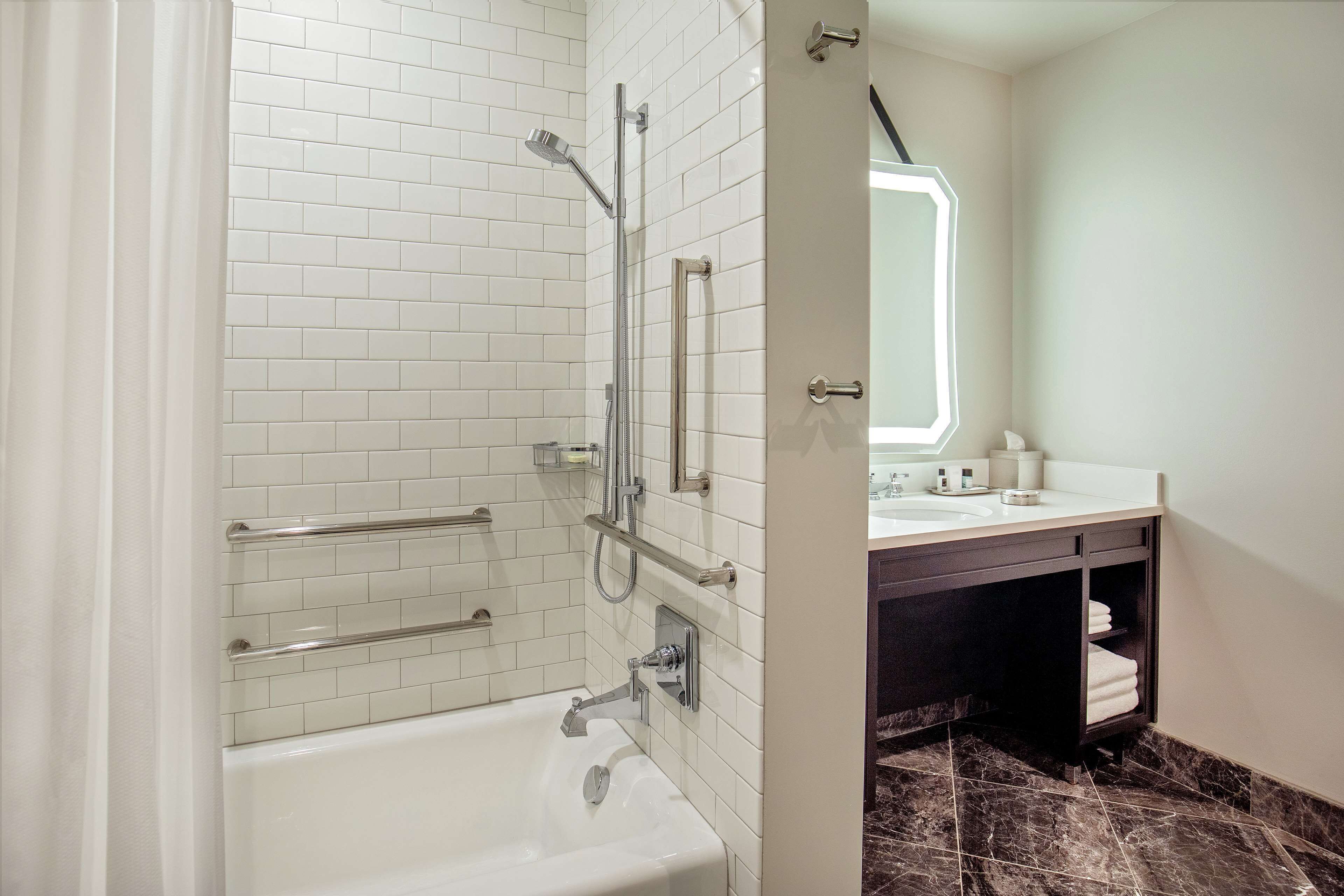 Un baño moderno con azulejos blancos tipo metro, una bañera blanca, ducha con mampara de vidrio y un mueble de tocador oscuro con espejo.