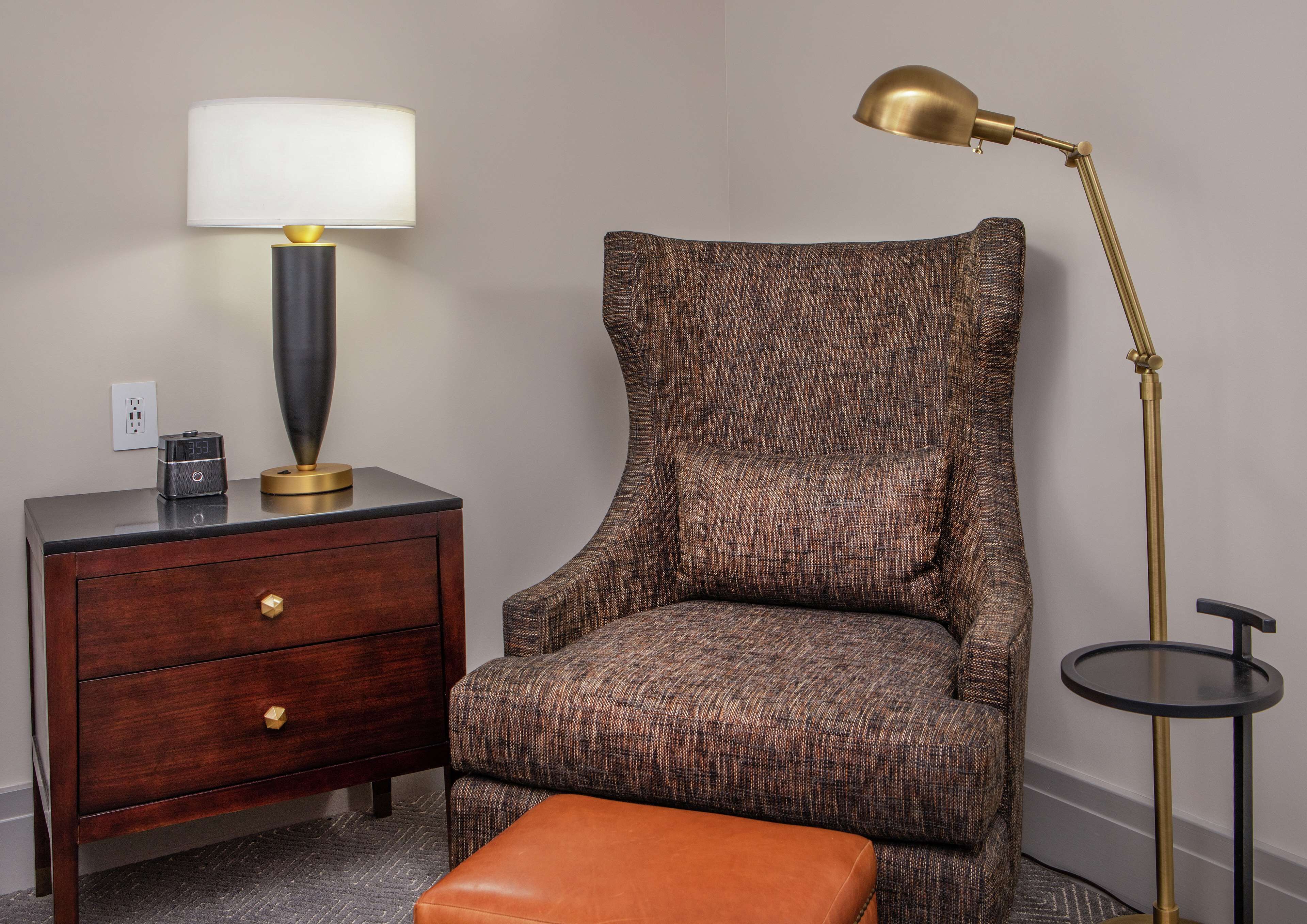 A bedroom with a wooden nightstand, a lamp, a chair, and a small round table.