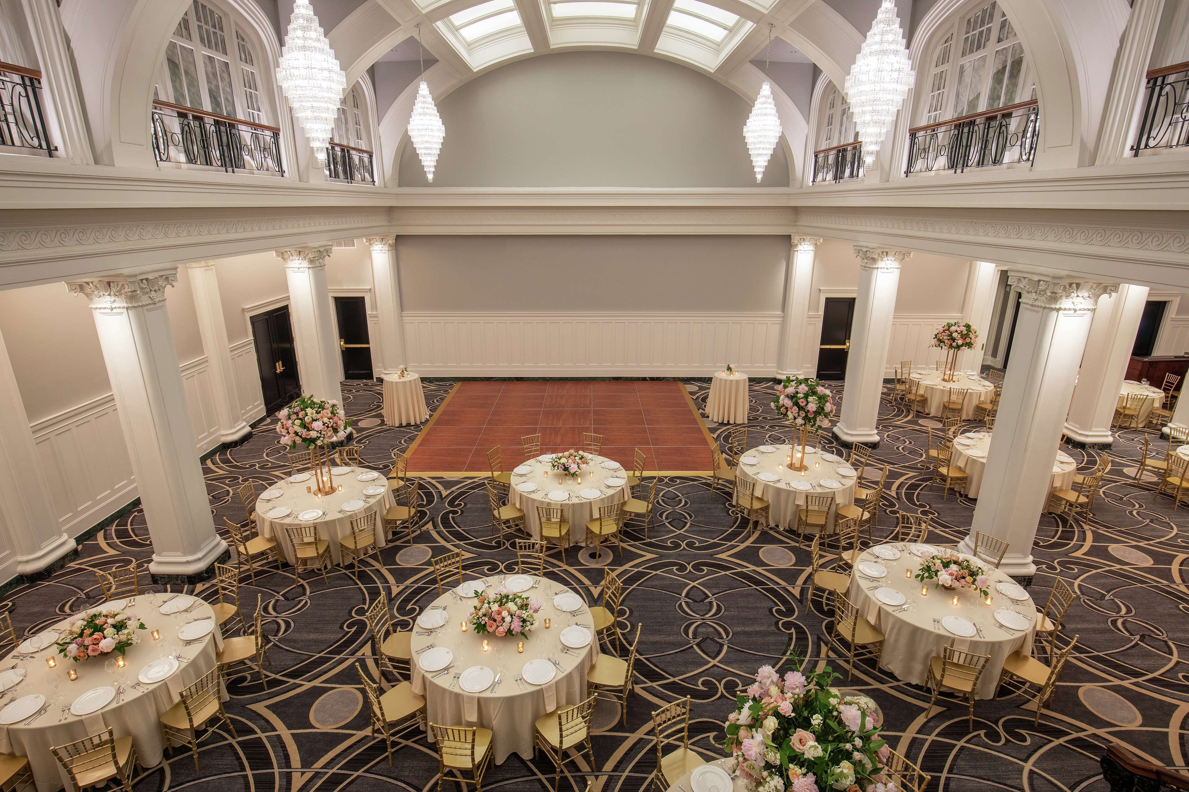 Ein prachtvoller Ballsaal mit mehreren runden Tischen, gedeckt mit weißen Tischdecken, goldenen Stühlen und floralen Mittelpunkten.