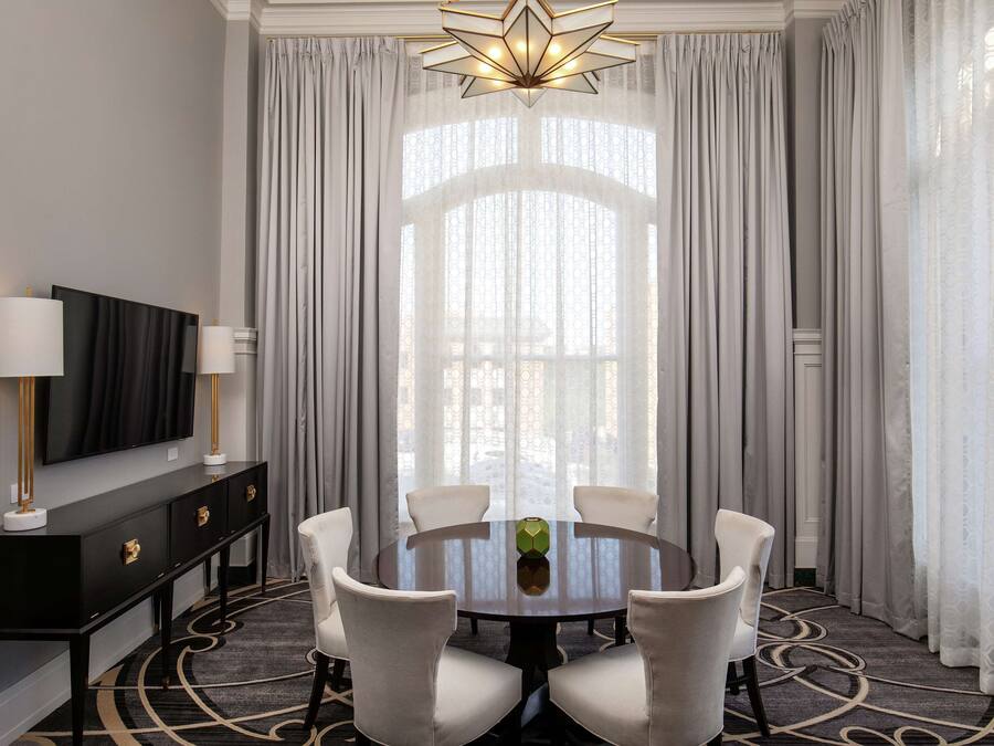 A modern dining area with a round table, six chairs, a black console table with a TV, and a large window with sheer curtains.
