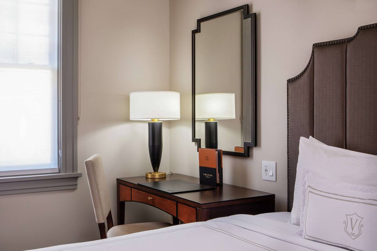 Een hotelkamer met een bed, een bureau met een lamp, een stoel, een grote spiegel en een raam.