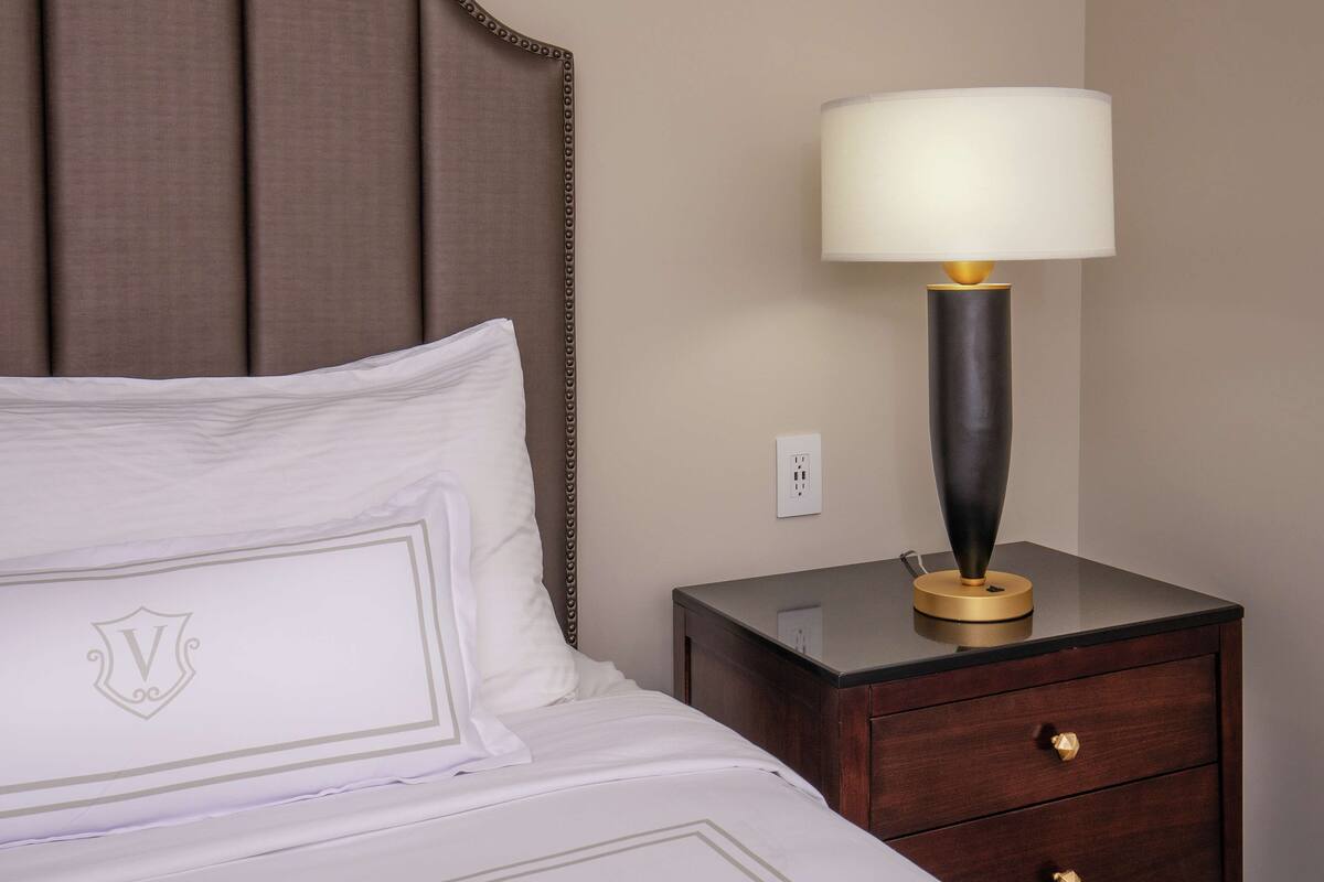 Een net opgemaakt bed met een witte deken en kussens, een nachtkastje met een lamp, en een stopcontact aan de muur.