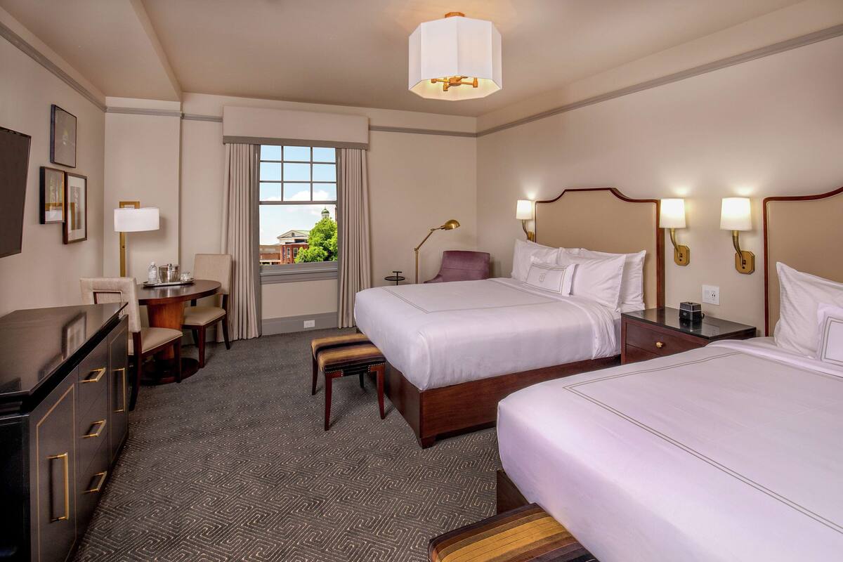 Een hotelkamer met twee bedden, een bureau, een stoel, een lamp en uitzicht naar buiten.