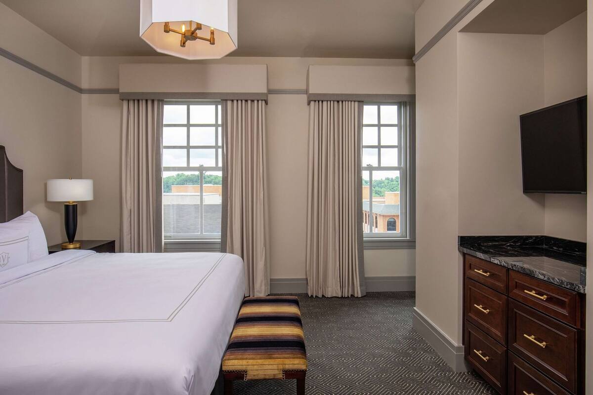 Een hotelkamer met een groot bed, een bureau met lamp, een bankje, een televisie en uitzicht op de stad door de ramen.