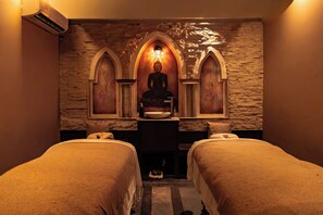 Una habitación con dos camas, una pared de piedra, una estatua y una pequeña mesa con un cuenco.