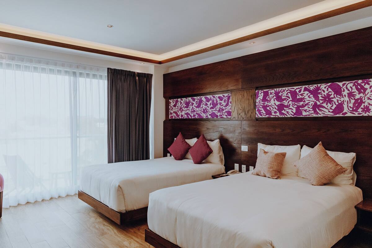 Ein Hotelzimmer mit zwei Betten, einem großen Fenster mit durchsichtigen Vorhängen und einem Holzkopfende mit dekorativer Zarge.