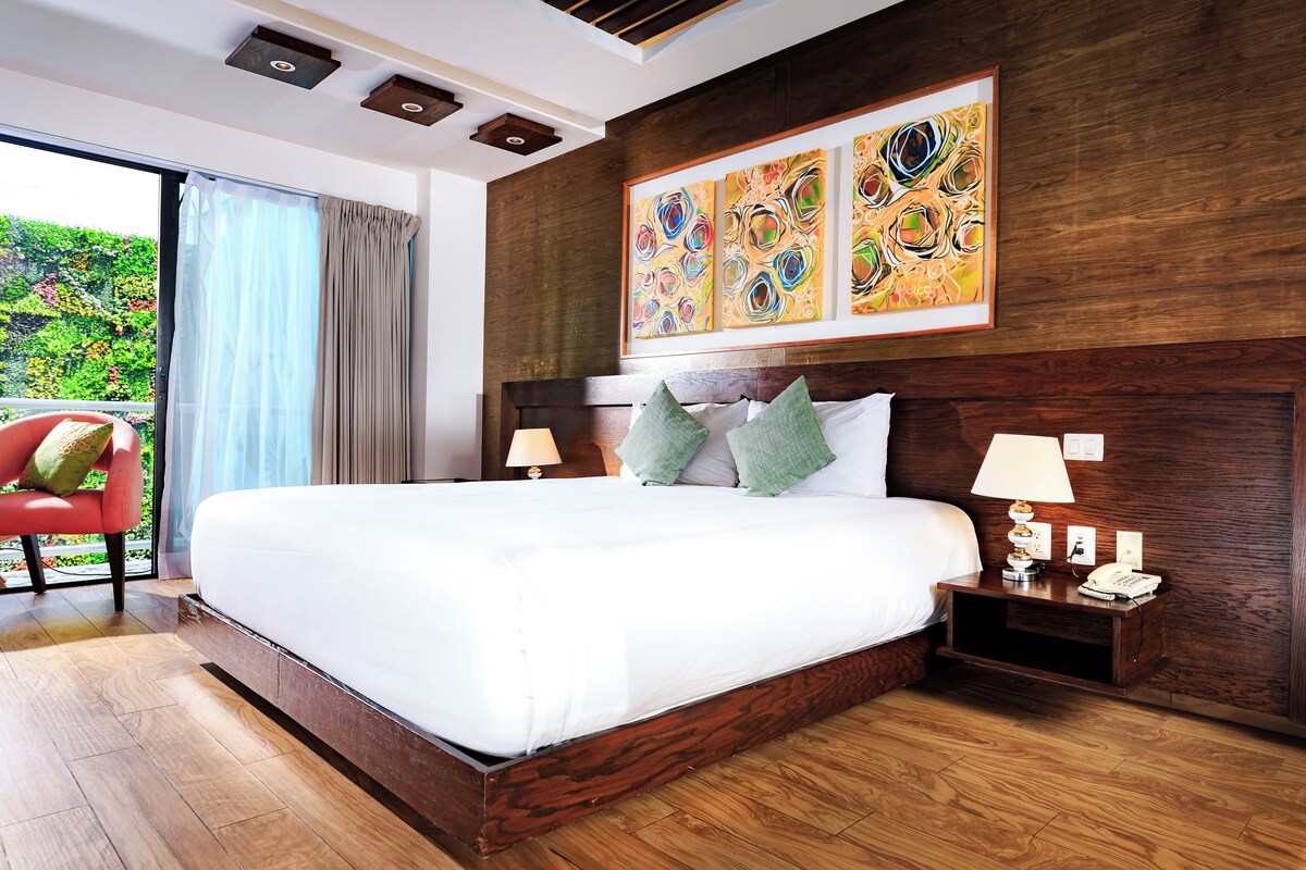 A modern hotel room with a large bed, wooden headboard, and a view of greenery through the window.