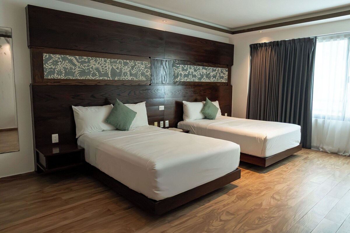 A hotel room with two beds, wooden flooring, and a large window with curtains.