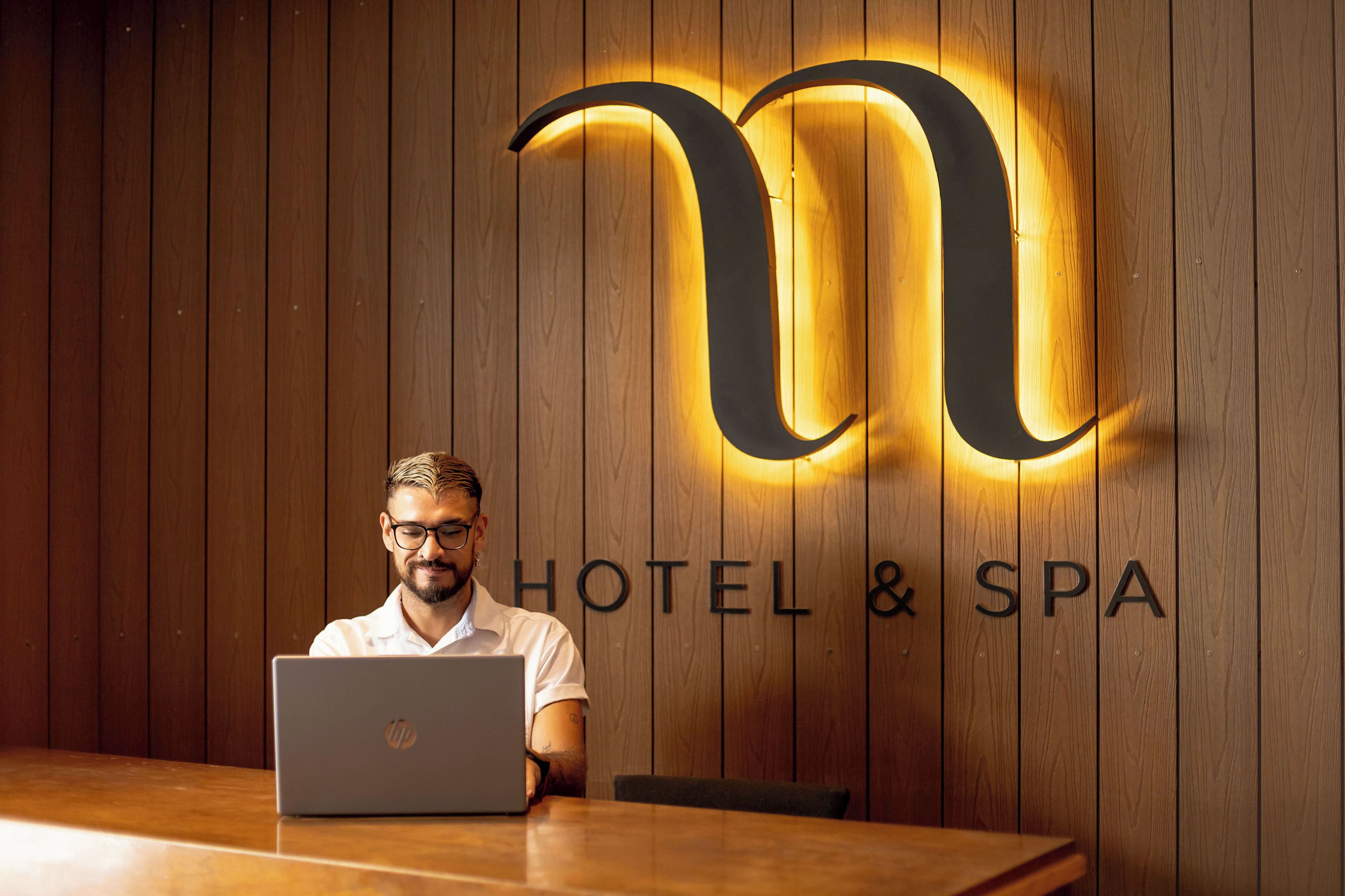 Un hombre sentado a una mesa de madera con una computadora portátil, frente a una pared con diseño de paneles de madera y una gran letra 'M' iluminada con el texto 'HOTEL & SPA' debajo.