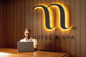 Un homme est assis à une table en bois, devant un ordinateur portable. Il est placé devant un mur orné de panneaux en bois et d’une grande lettre « M » illuminée, avec le texte « HÔTEL ET SPA » en dessous.