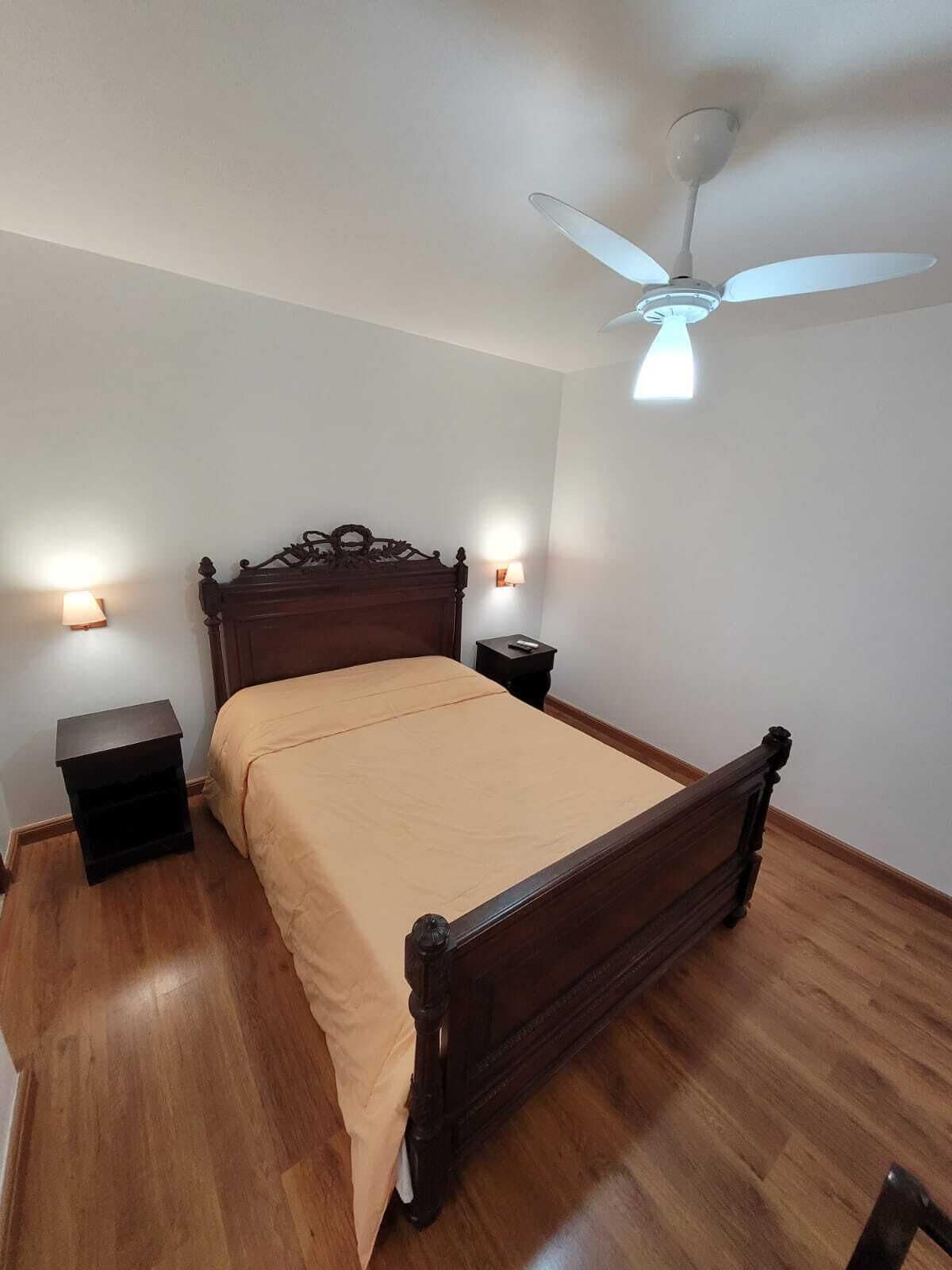 Quarto com cama de madeira, duas mesas de cabeceira, ventilador de teto e luminária na parede.