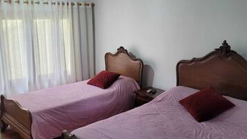 A room with two wooden beds, each with a headboard and a red pillow. There is a window with white curtains and a wooden ceiling.