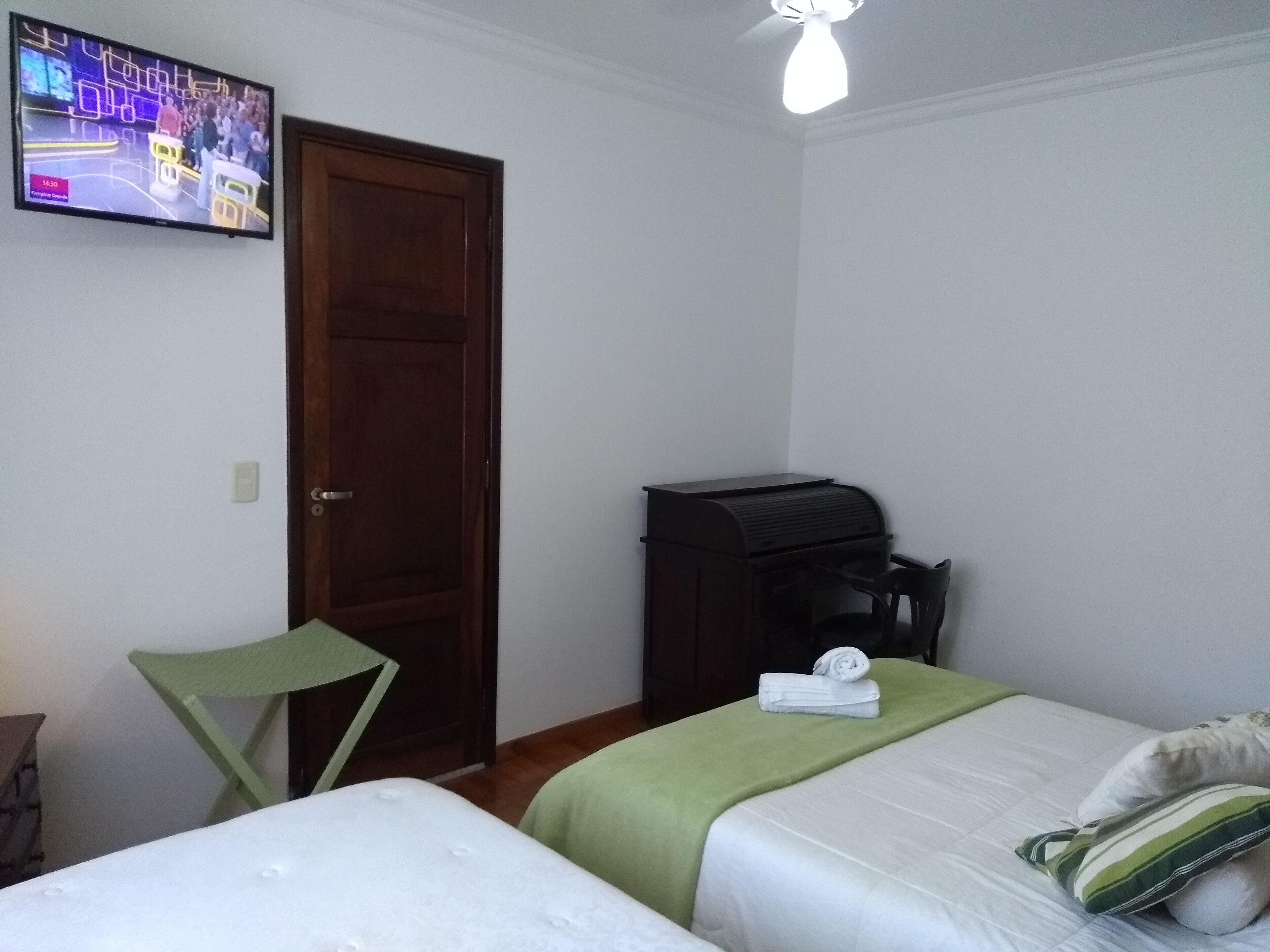 Quarto de hotel com duas camas, uma televisão na parede, uma porta de madeira e uma pequena mesa com uma chaleira.