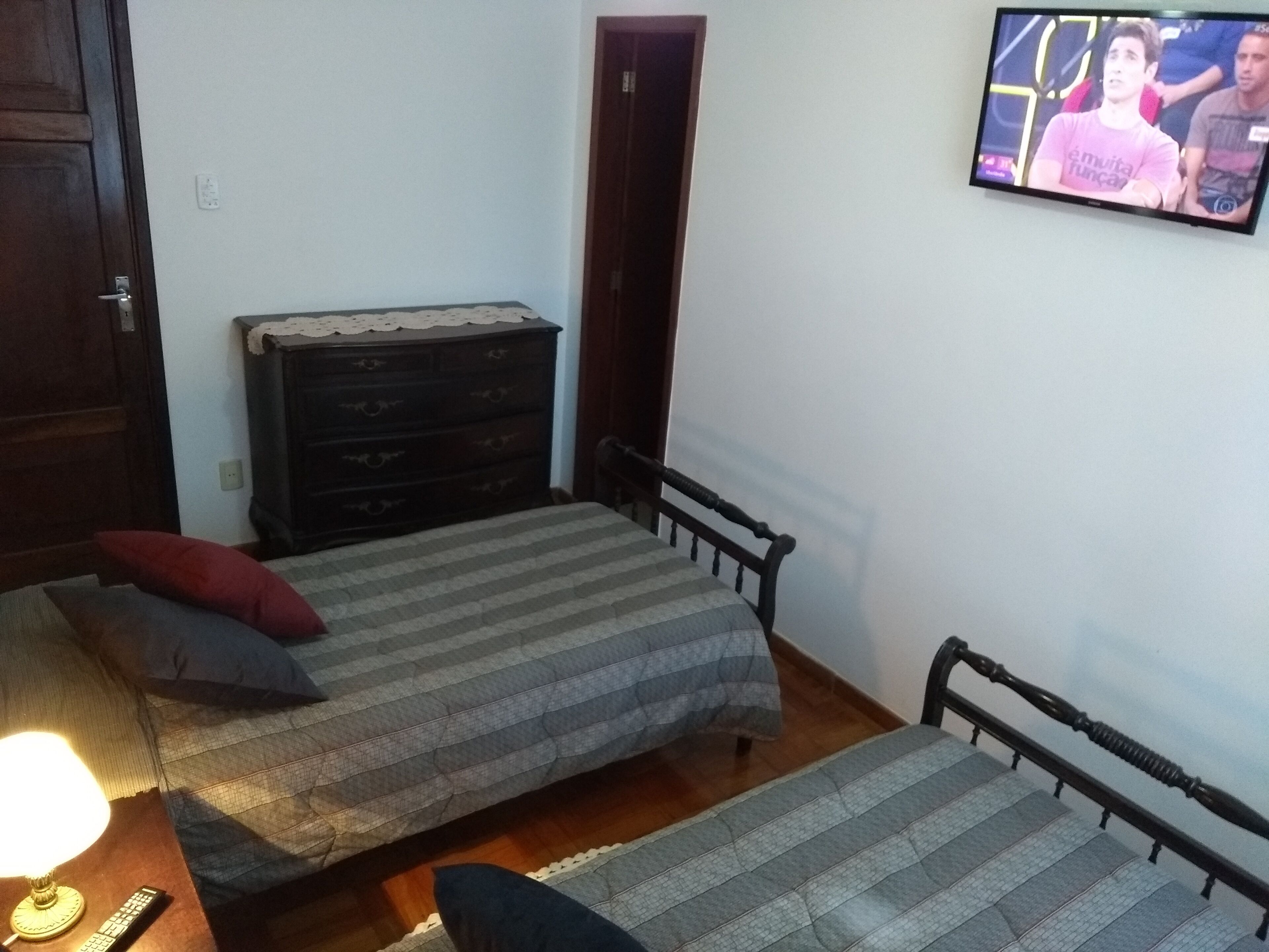 A small, simple room with two single beds, a dresser, a television mounted on the wall, and a door.
