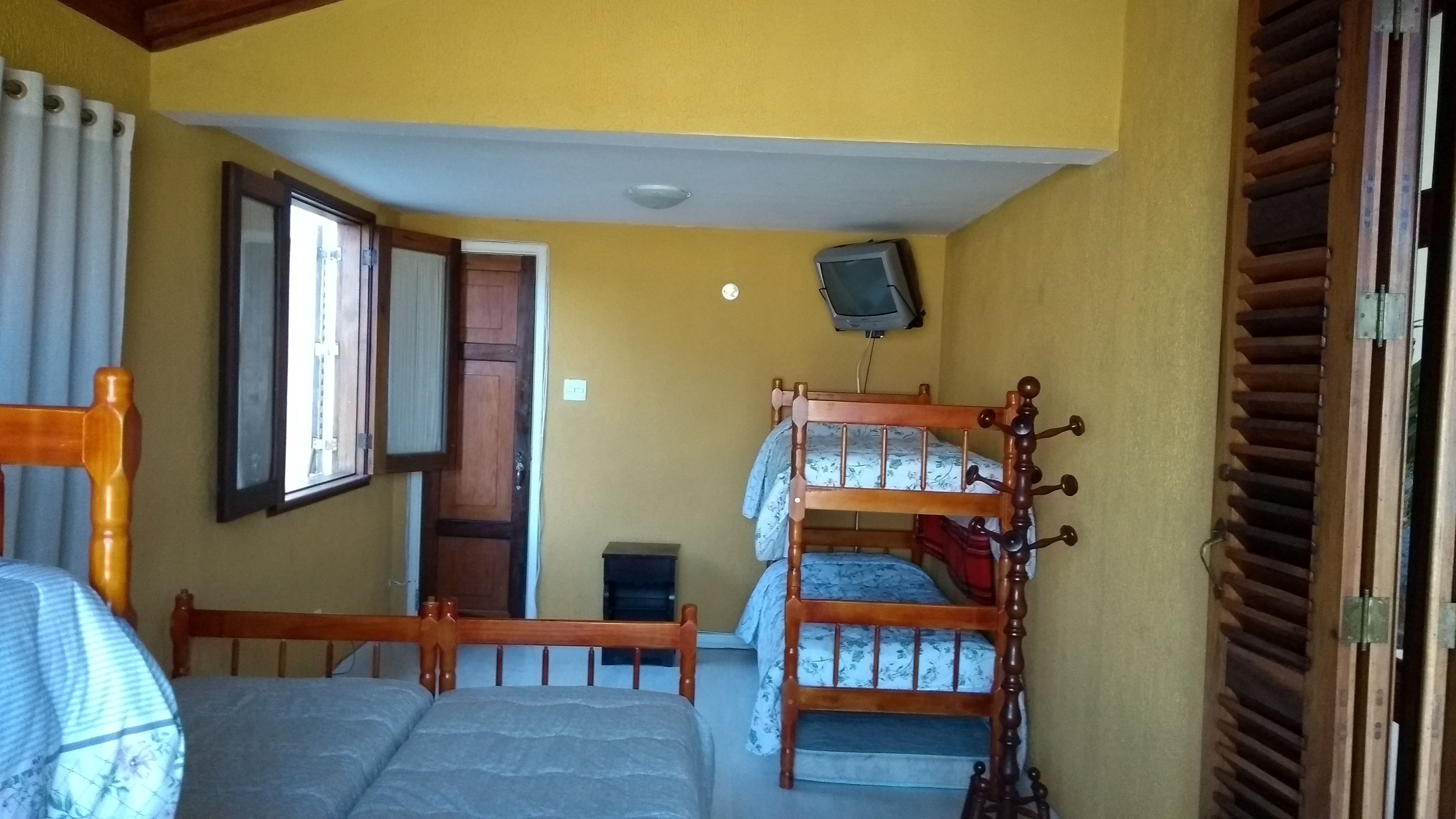 Quarto com duas beliches de madeira, uma televisão presa à parede e uma janela com cortinas.