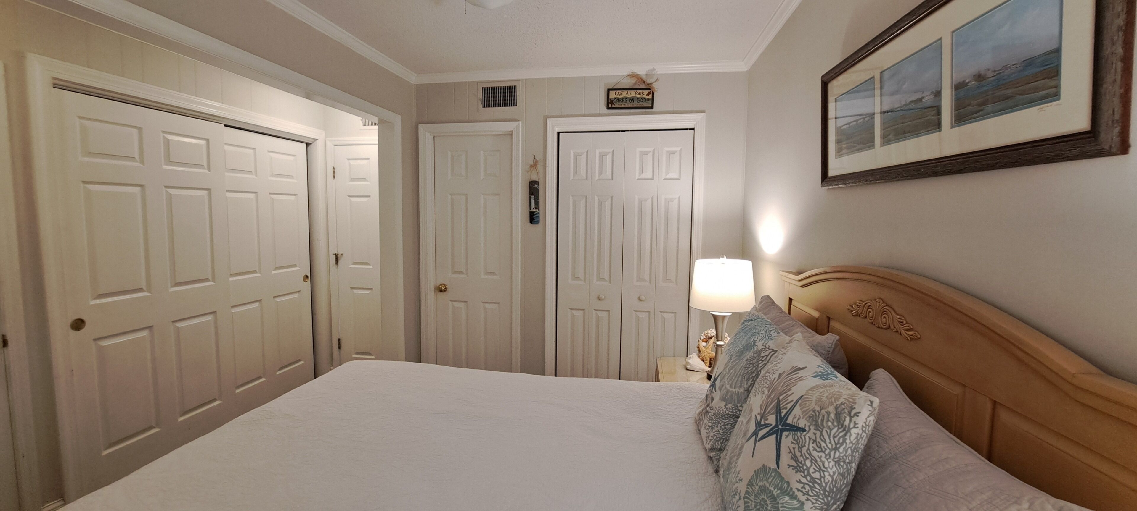 A hotel room with a bed, a nightstand with a lamp, a framed picture on the wall, and two doors.