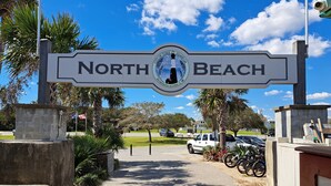 Un panneau indiquant l’accès à North Beach, orné d’un emblème représentant un phare, et entouré de palmiers et d’une promenade dallée.