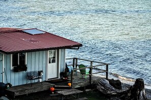 A small cabin with a red roof, a dock extending into the water, and a chair on the dock.