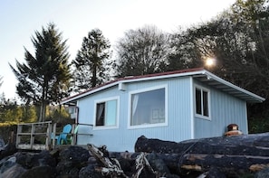 Une petite cabane bleu clair, dotée de deux fenêtres et d’un toit rouge, entourée d’arbres et de troncs d’arbres.