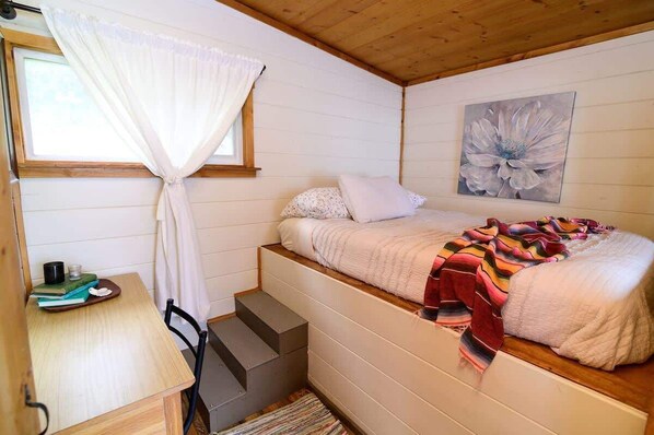 A bedroom with a bed, a window with curtains, a painting on the wall, and a small table with books and a cup.