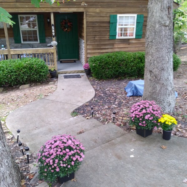 A wooden house with a green door, a mailbox, and a number '589' on the wall. There are potted plants with pink and yellow flowers in front of the house. - Cozy Cabin Located in gated resort near Lake Cumberland. Pool & boat ramp onsite (Burnside)