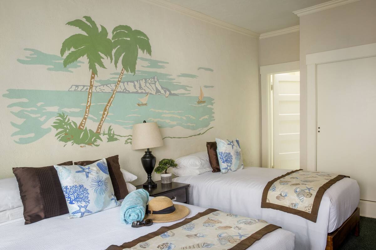 Habitación de hotel con dos camas, una mesita de noche con lámpara y un mural en la pared con una playa.