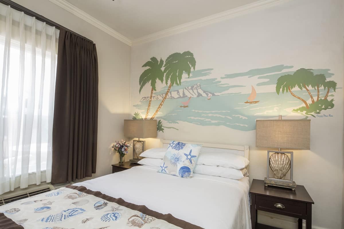 Habitación de hotel con cama, mesita de noche, lámpara, un mural en la pared con una escena de playa y una ventana con cortinas.