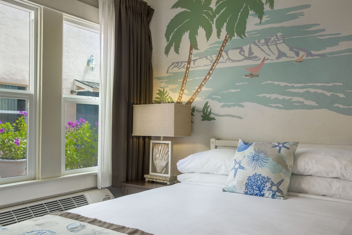 Una habitación de hotel con una cama, una mesita de noche con una lámpara, una ventana con cortinas y un mural de playa en la pared.