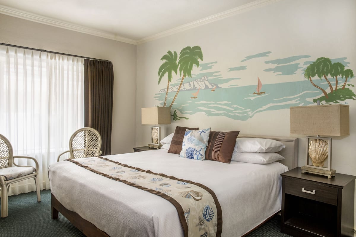 Una cama bien hecha con un mural temático de playa, una silla y una mesita de noche con lámpara.
