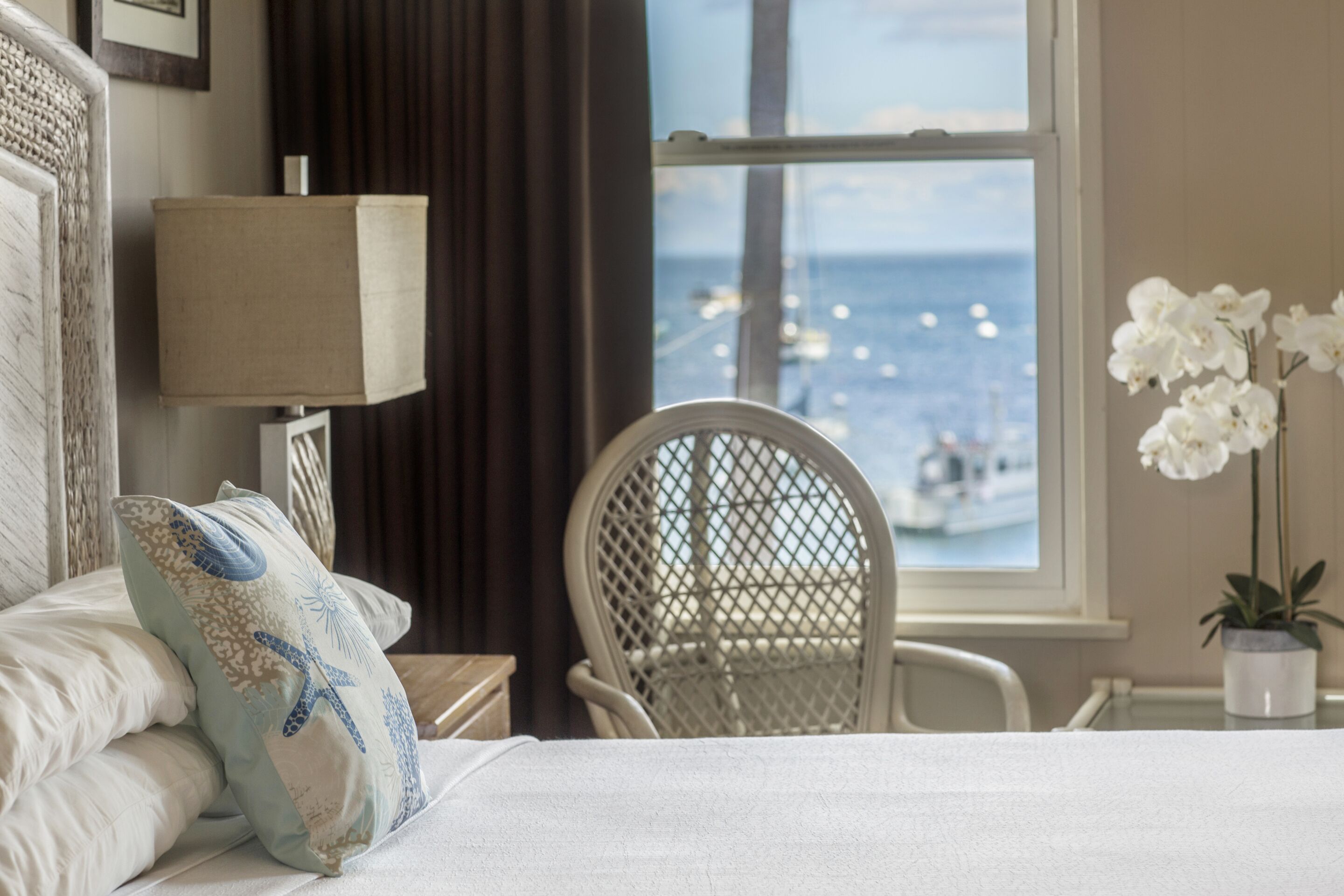 A hotel room with a bed, a chair, a window with a view of the sea, and a potted plant.
