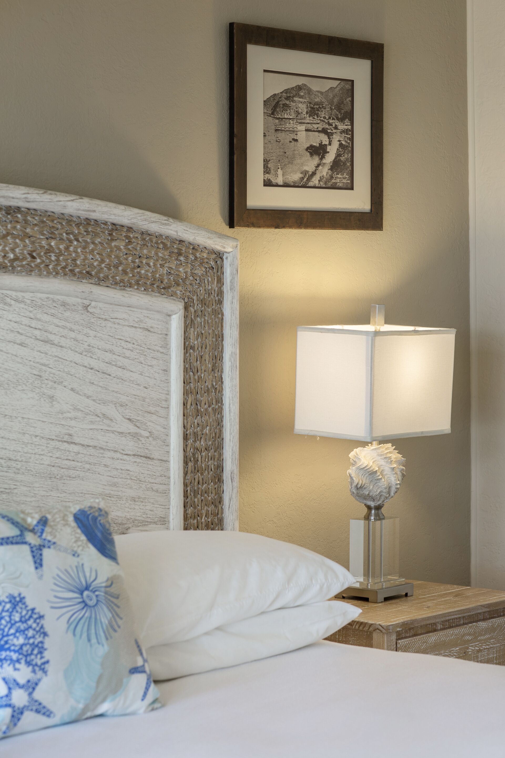 A neatly made bed with a white pillow and a blue floral pillow, a bedside lamp with a decorative base, and a framed black and white photograph on the wall.