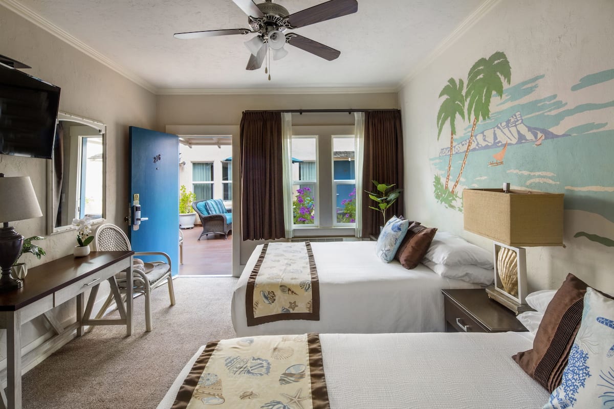 Habitación de hotel con dos camas, un escritorio, un televisor, un ventilador de techo y vistas al balcón exterior.